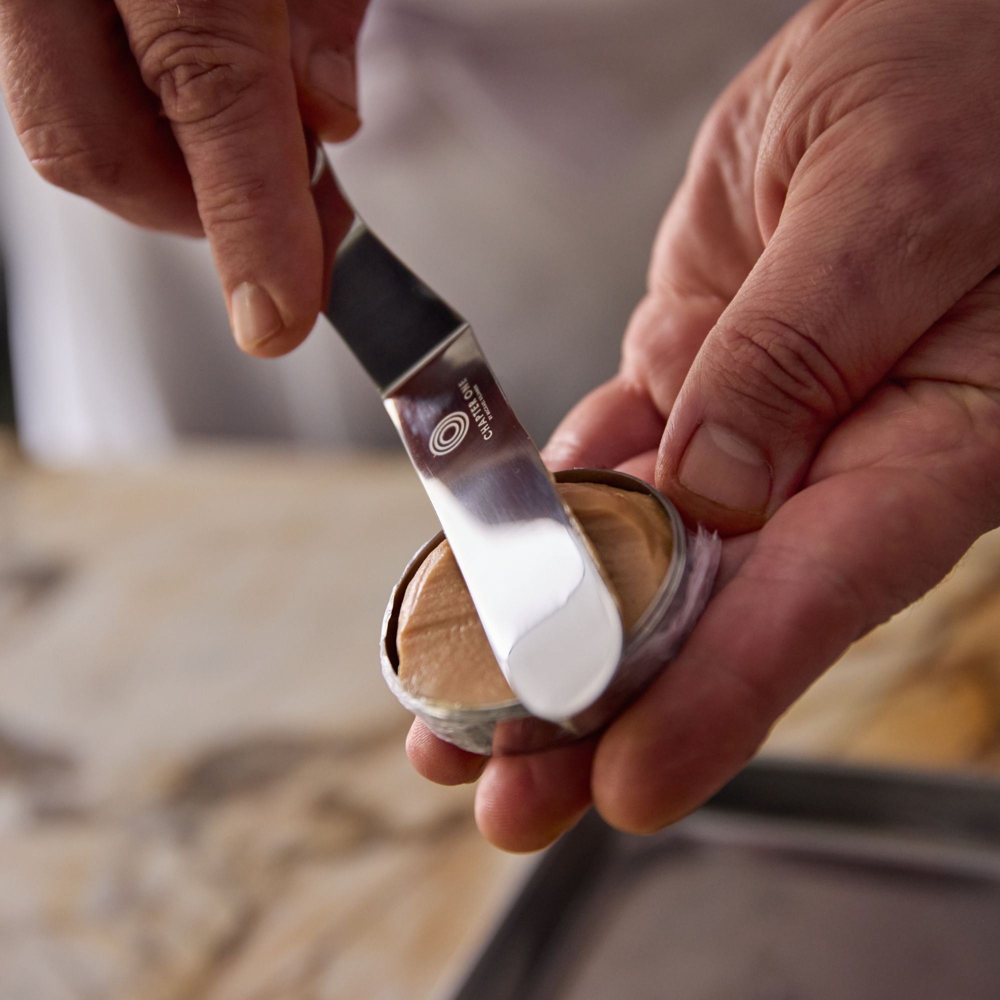 A close up of a Chef smoothing a tin of pate with a stainless steel palette knife that has an edge-to-edge sharp blade and the Chapter One Logo engraved on it.
