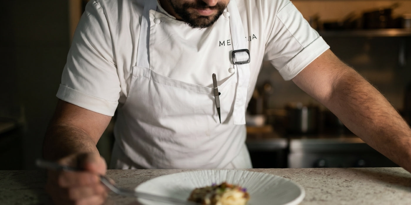 A chef leaning over a dish while plating with small kitchen tweezers secured to a magnetic pin attached to their apron, keeping the tweezers easily accessible during food preparation.