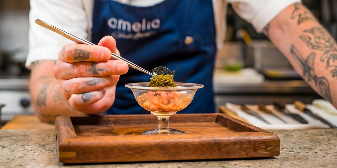 A chef wearing a blue apron uses precision tweezers to arrange ingredients in a clear glass bowl, focusing on the final  presentation of the dish.