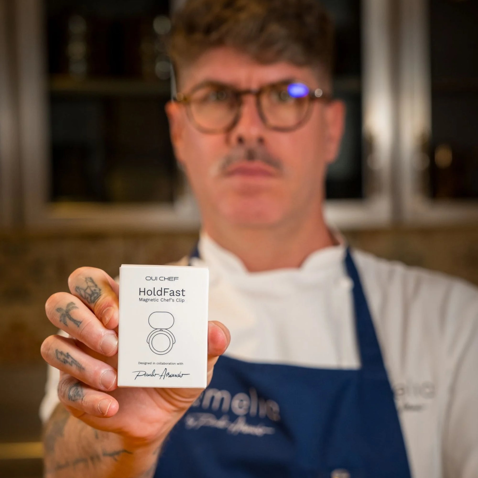 Chef wearing a blue apron, in a professional kitchen holding the 'HoldFast Magnetic Chef's Clip' packaging
