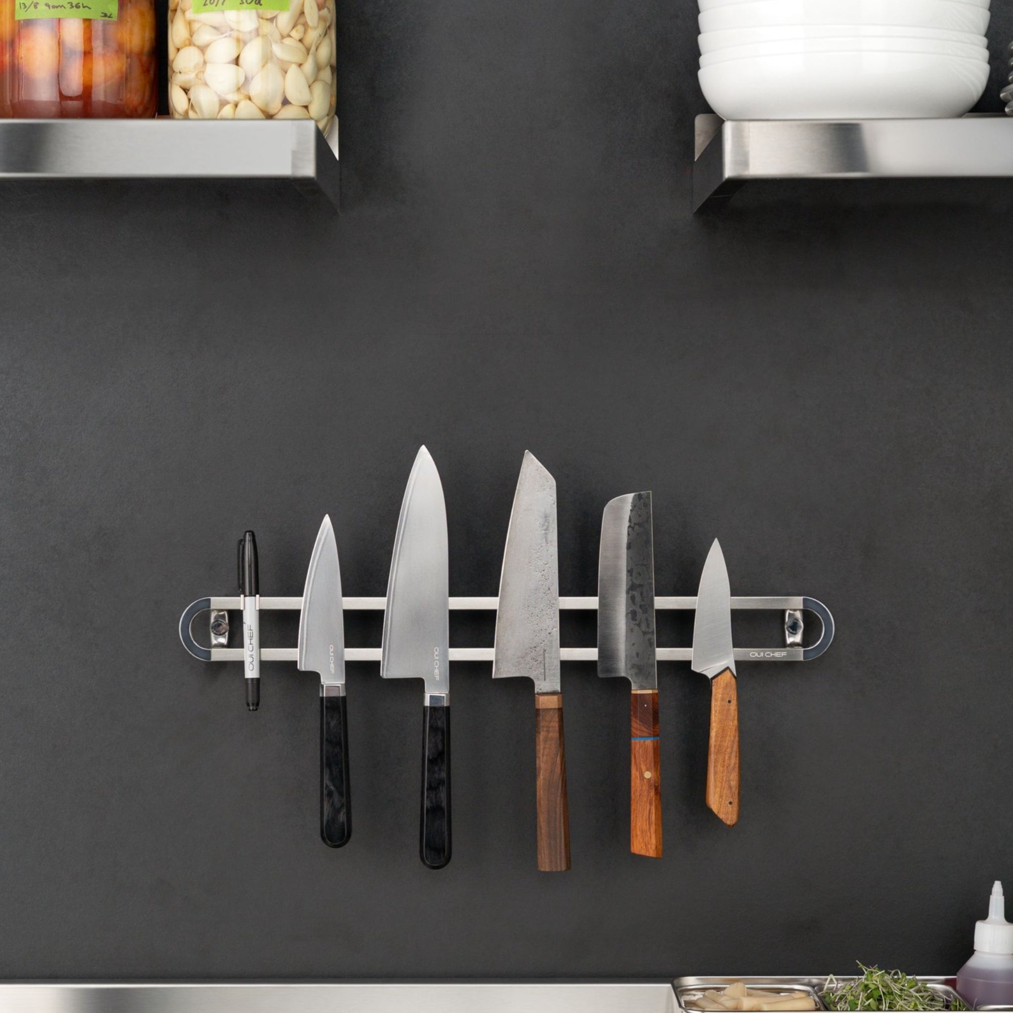 Stainless steel magnetic rack placed on a charcoal coloured restaurant kitchen wall with five chef's knives on it and a sharpie attached to a clip.  
